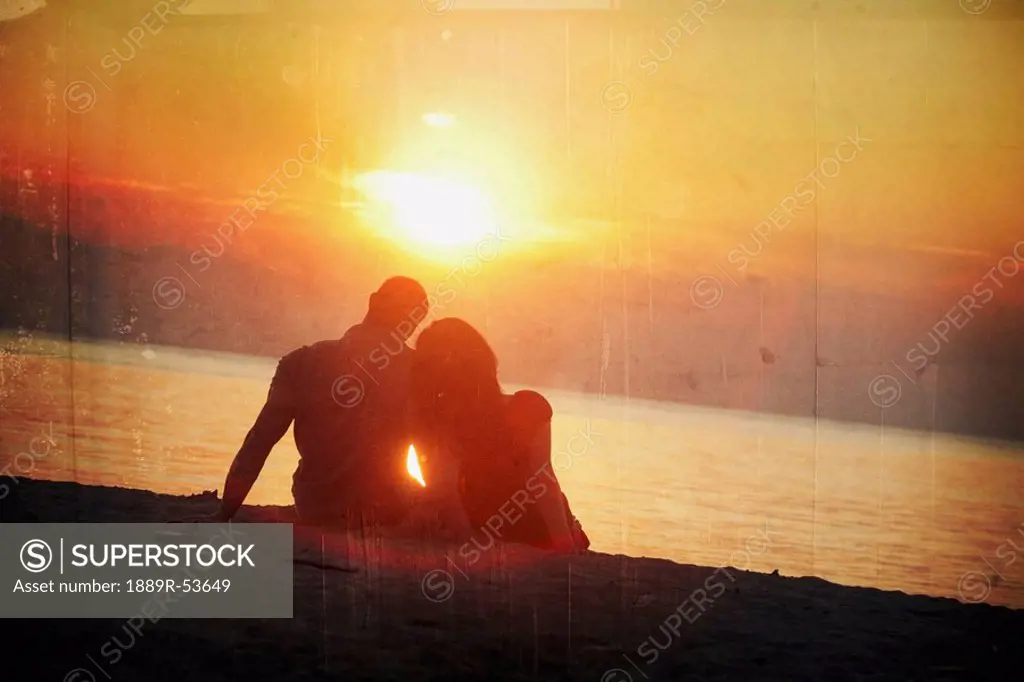 a man and woman sitting on the beach in a sunset