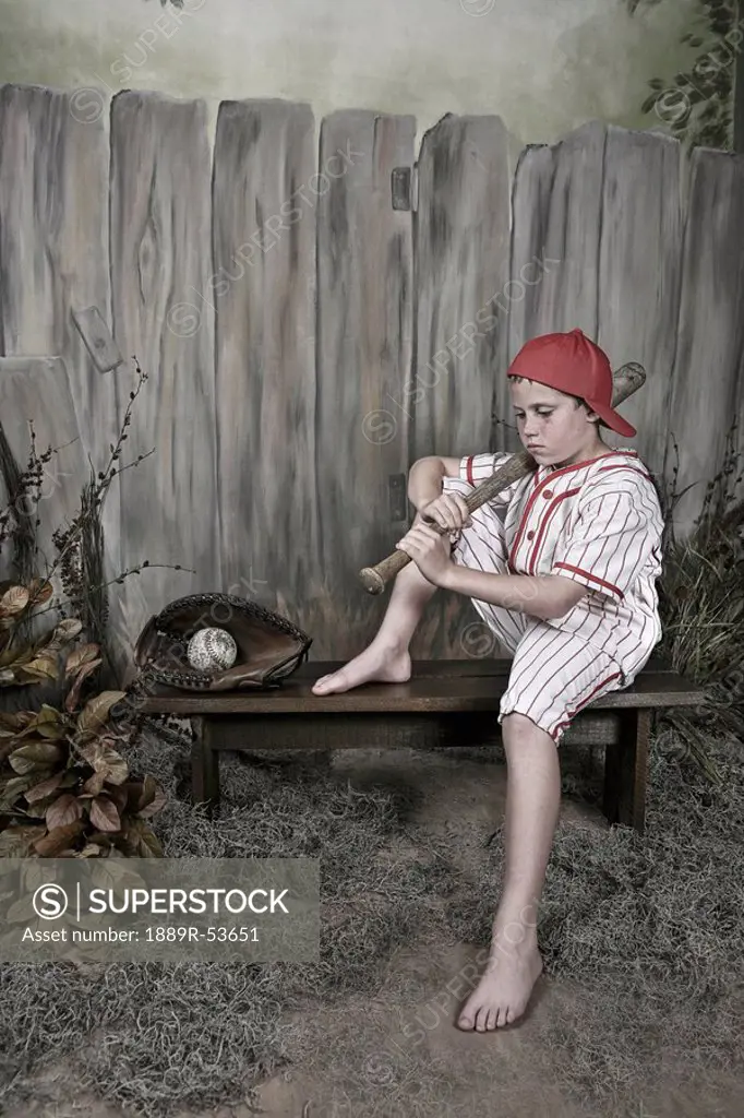 a boy holding a baseball bat and wearing a uniform sitting on a bench