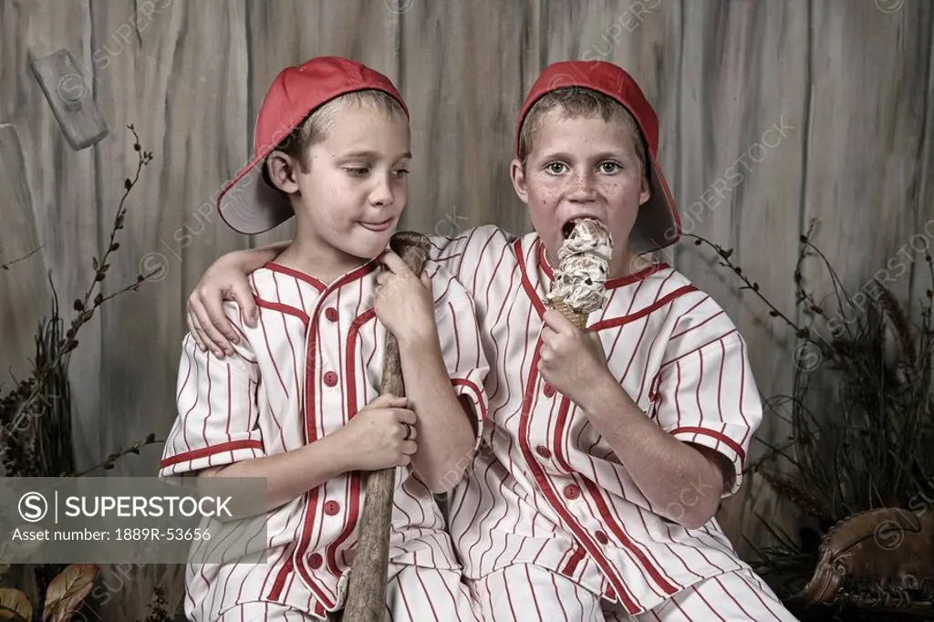 two boys wearing baseball uniforms and one is eating an ice cream cone