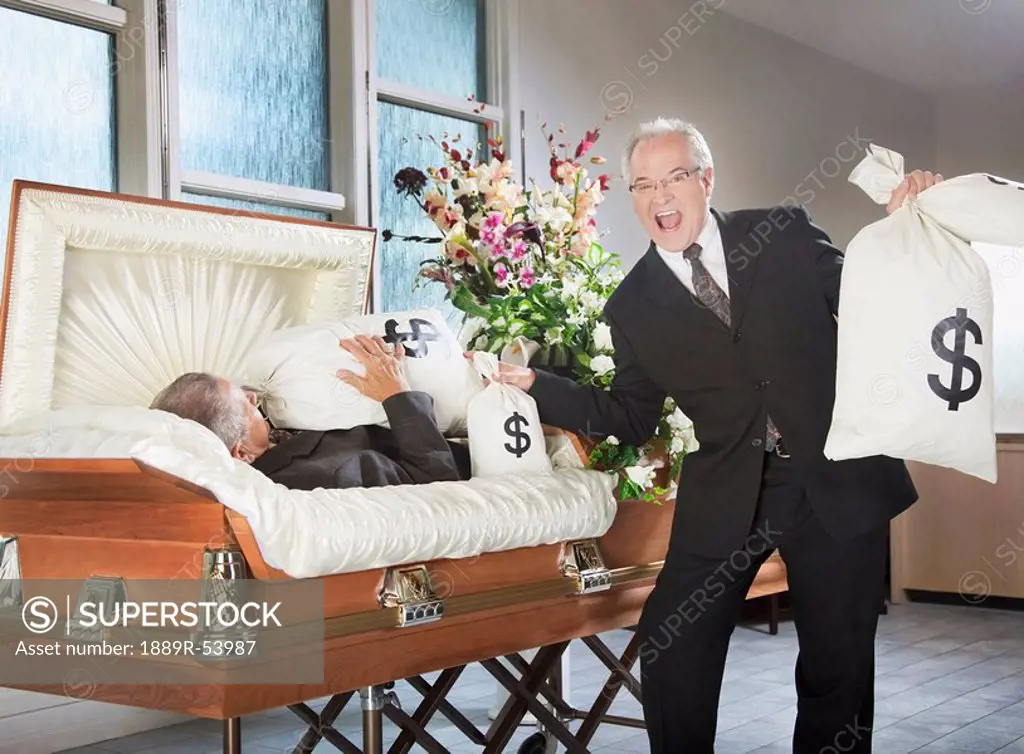 edmonton, alberta, canada, a happy man holds bags of money beside a deceased man in a coffin