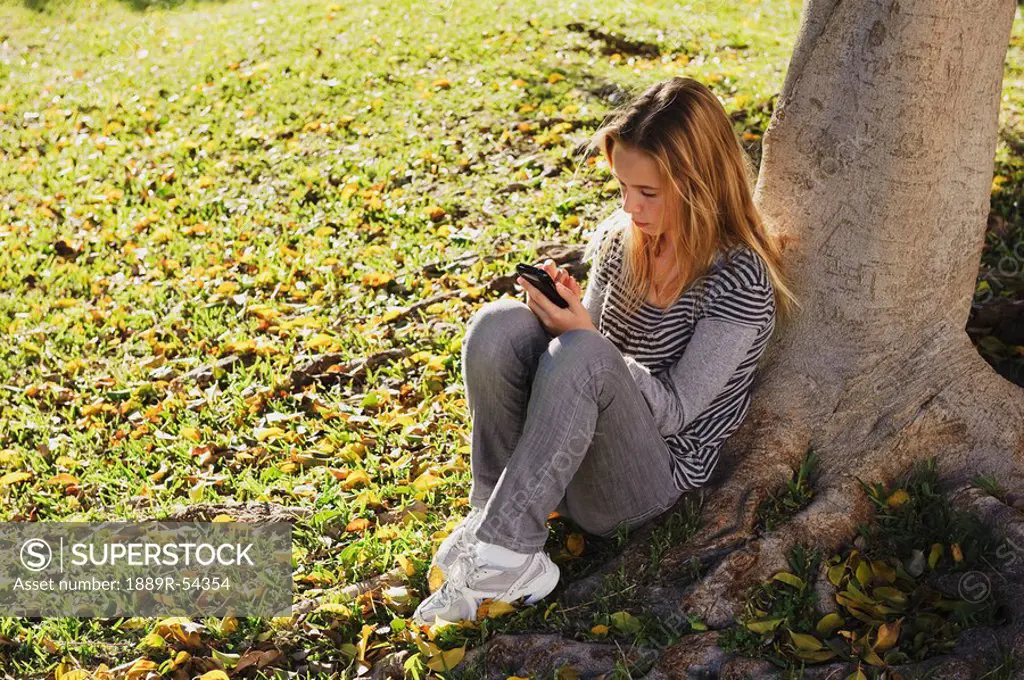 a young girl sits under a tree at the park using her cell phone