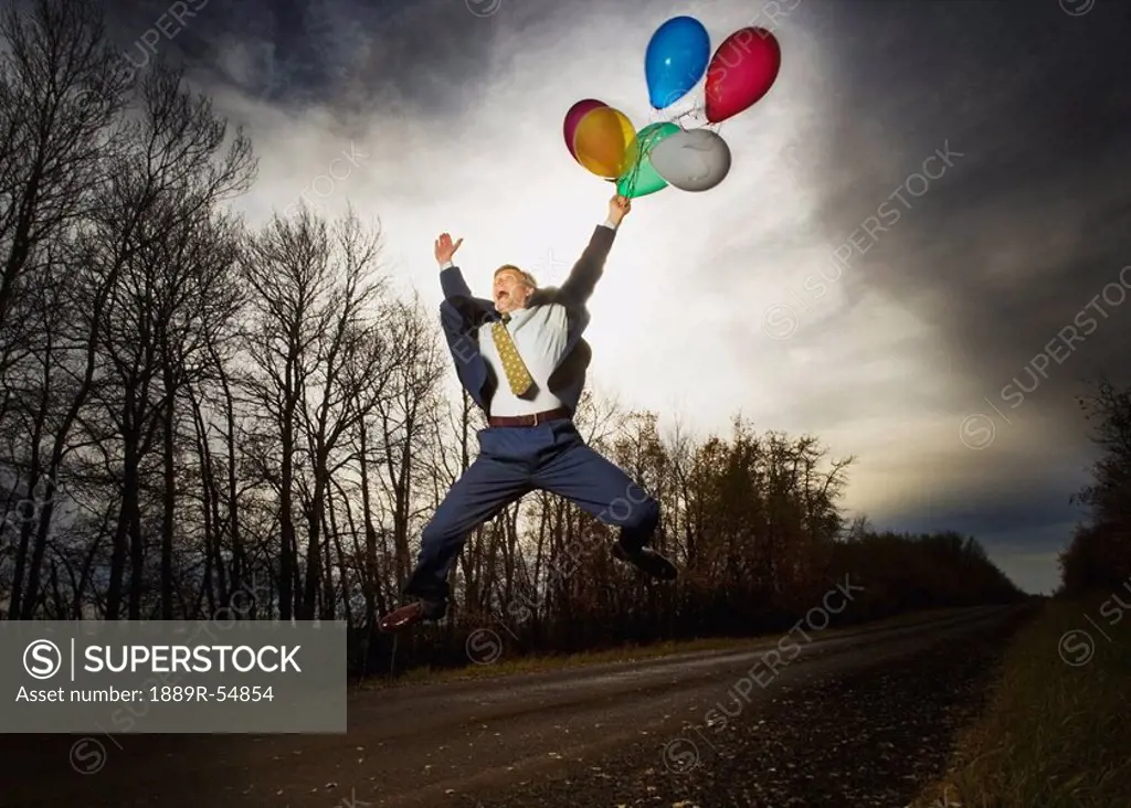 Alberta, Canada, A Businessman Jumping In The Air On The Side Of The Road Holding Balloons