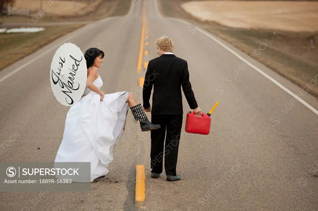 three hills, alberta, canada, a bride and groom walk down a rural road with a jerry can and a parasol saying ´just married´