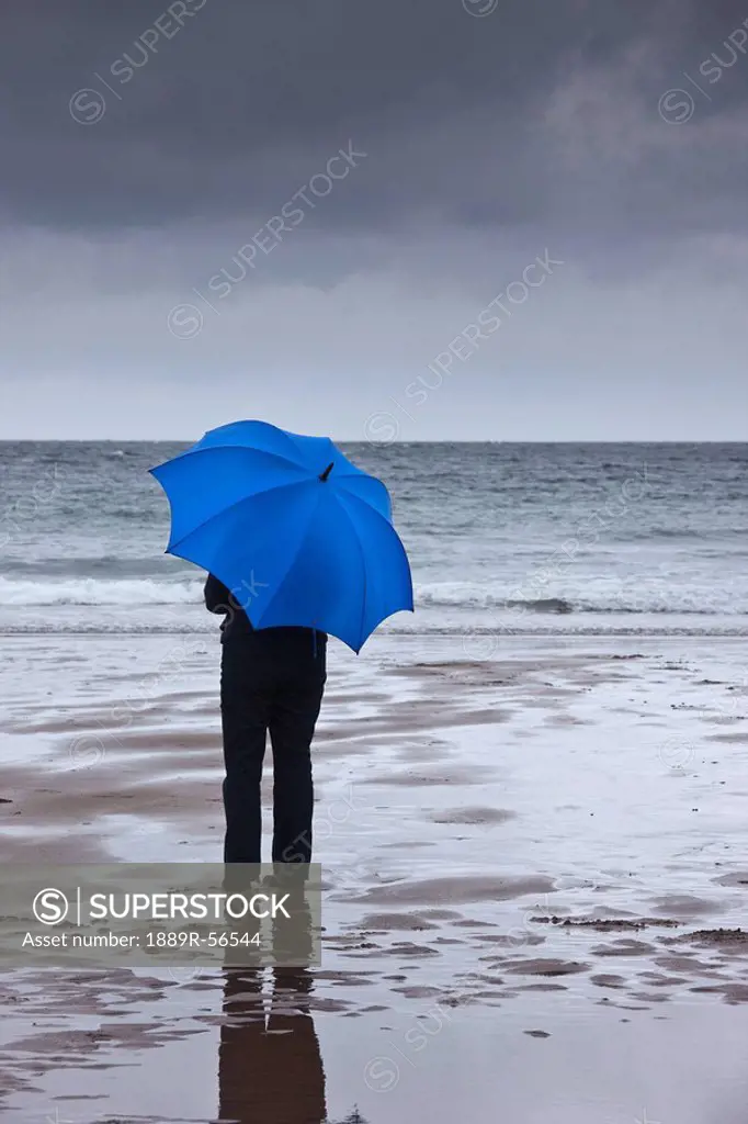 a person holding a blue umbrella standing on the beach under a dark sky, northumberland, england
