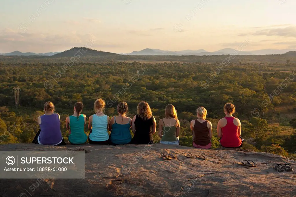 A Row Of Young Women Sitting On A Rock Ledge Viewing The African Landscape, Manica, Mozambique, Africa