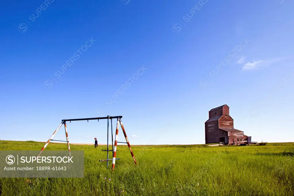 A Man Walking Through A Field With An Old Swing Set And An Old Grain Elevator, Bents, Saskatchewan, Canada