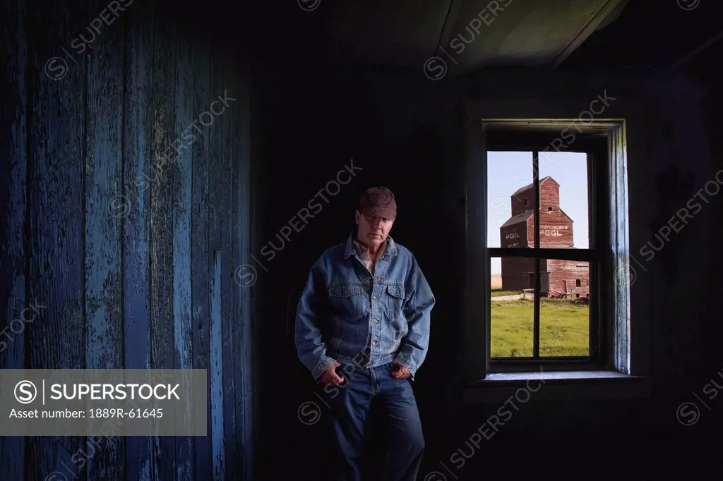 Portrait Of A Farmer/Rancher In An Old Abandoned Ghost Town Store With A Grain Elevator Seen Out The Window, Bents, Saskatchewan, Canada
