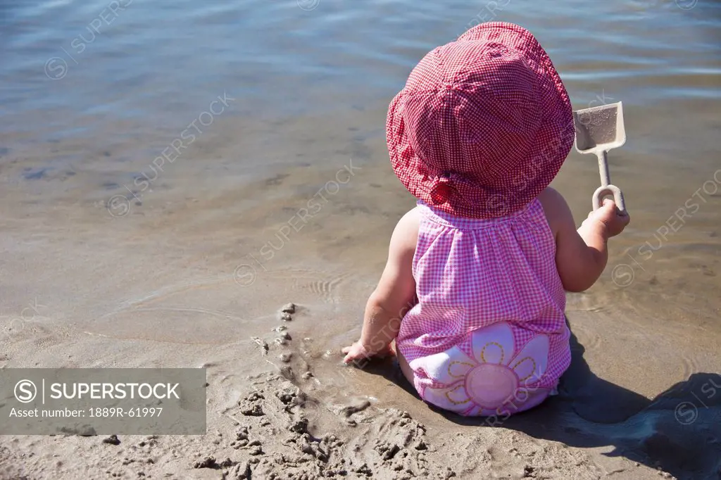 a young girl sits in the wet sand at the water´s edge holding a shovel, county of st. paul, alberta, canada