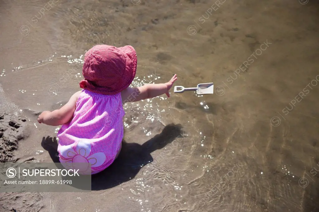 a young girl sits in the wet sand at the water´s edge reaching for her floating shovel, county of st. paul, alberta, canada