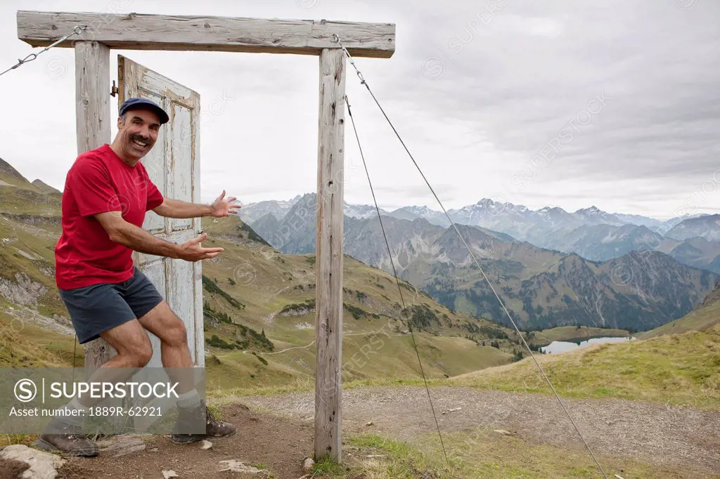 male hiker on top of a mountain stepping into an open wooden door and frame overlooking a mountain valley, lake and mountain range, oberstdorf, german...