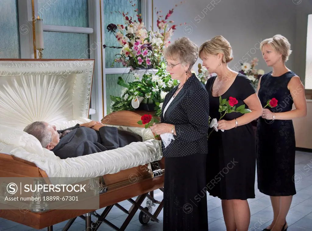three women standing by a man´s body laying in a coffin