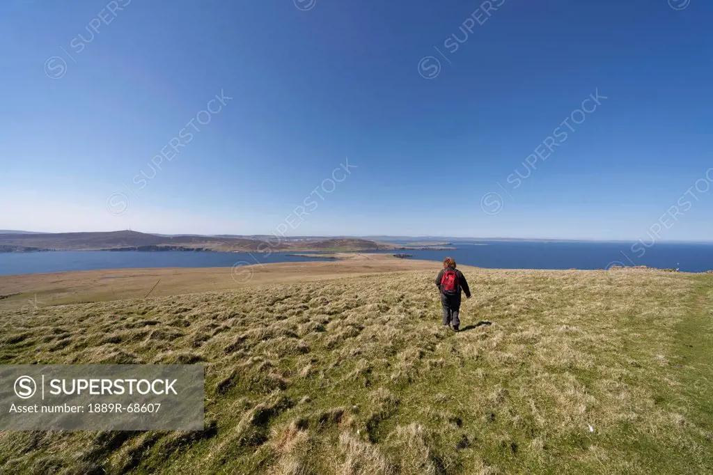 a woman walking across a grass field along the coast, noss scotland