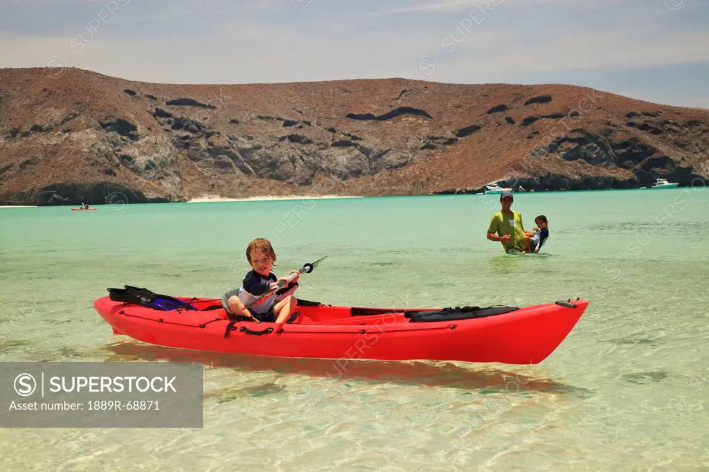 a young boy sits in a red boat by the shore while a father and son play out in the water at los islotes national marine park espiritu santo island, la...