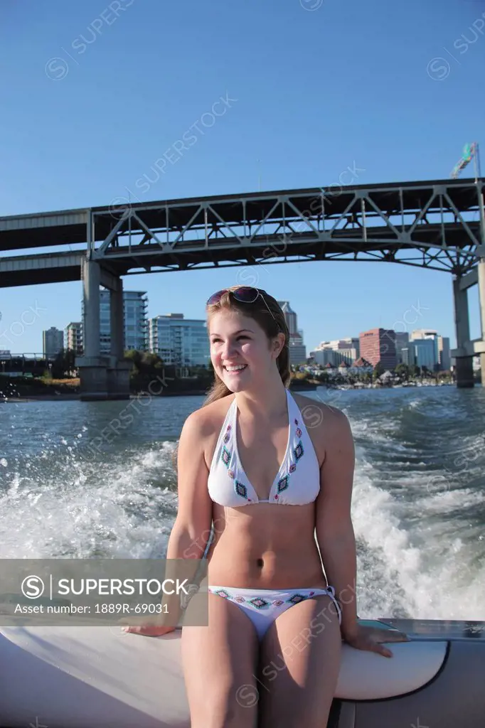 a teenage girl in a bathing suit sitting at the back of a boat with the city and a bridge in the background, portland oregon united states of america