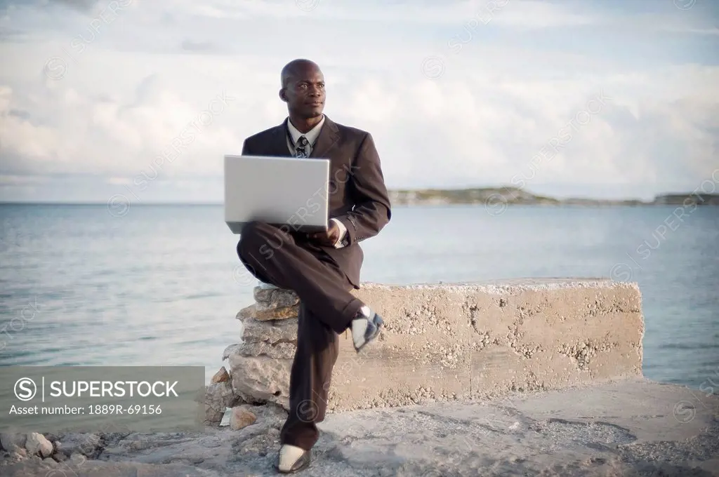 a businessman with a laptop computer sitting on a rock ledge at the edge of the water, south caicos turks and caicos islands