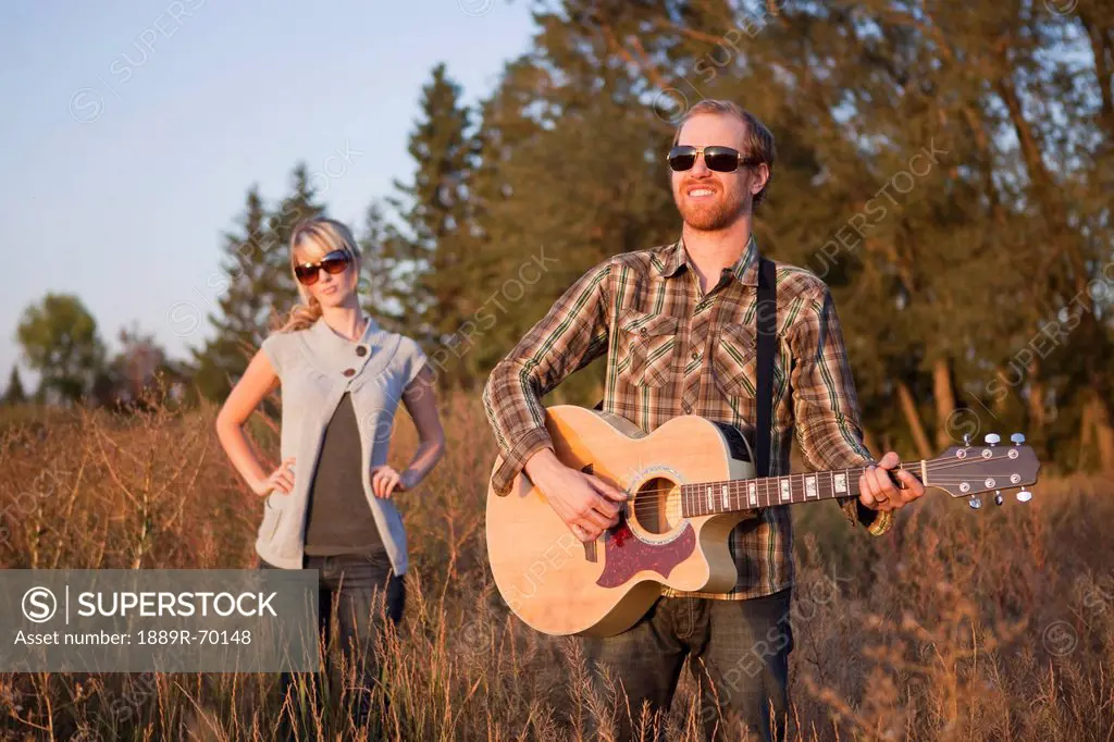 a young woman being ignored while standing in a field with a young man playing a guitar, edmonton alberta canada