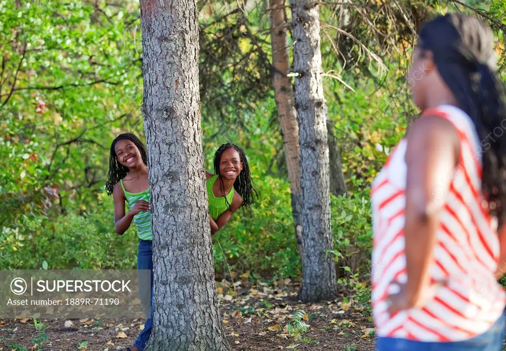 sisters playing hide and seek in a park with their mother looking on, edmonton alberta canada