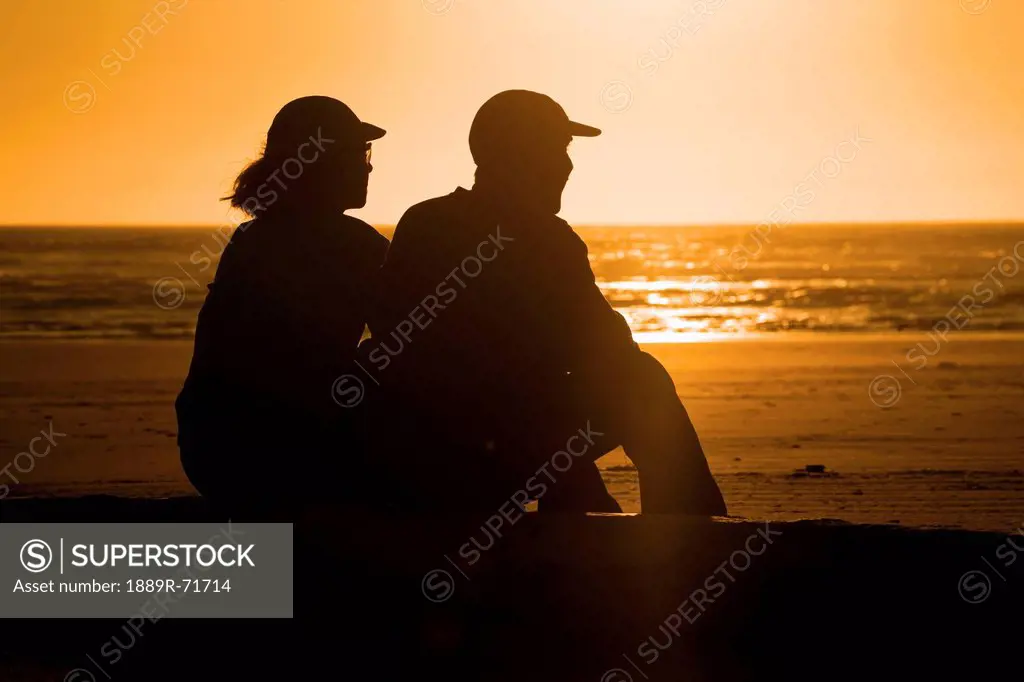 a silhouette of a couple sitting on a log on the beach at sunset, newport, oregon, united states of america