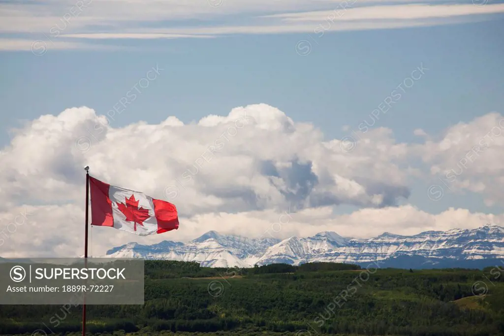 canadian flag blowing in the wind with snowy mountains in the distance and clouds with blue sky, alberta canada