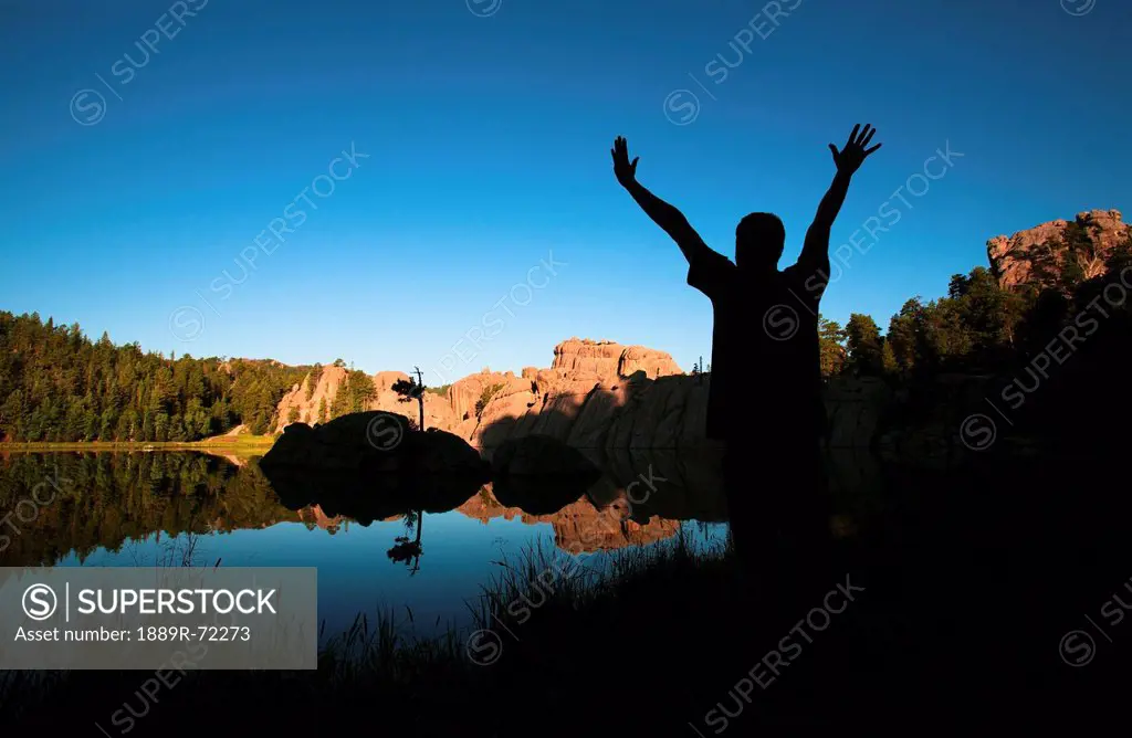 silhouette of a young man standing at the water´s edge of sylvan lake with his arm´s raised, black hills south dakota united states of america