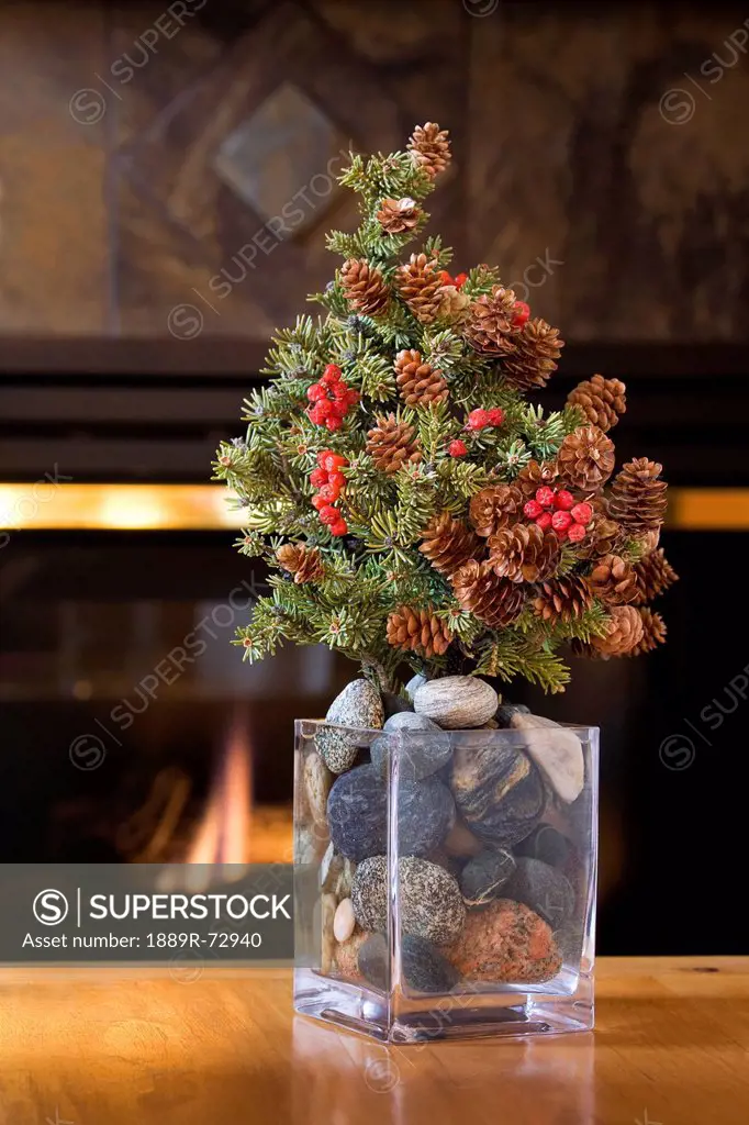 Small Real Fur Tree Decorated With Cones And Red Berries Christmas Tree On A Table In Glass Vase Filled With Round Stones And A Glowing Firplace In Th...
