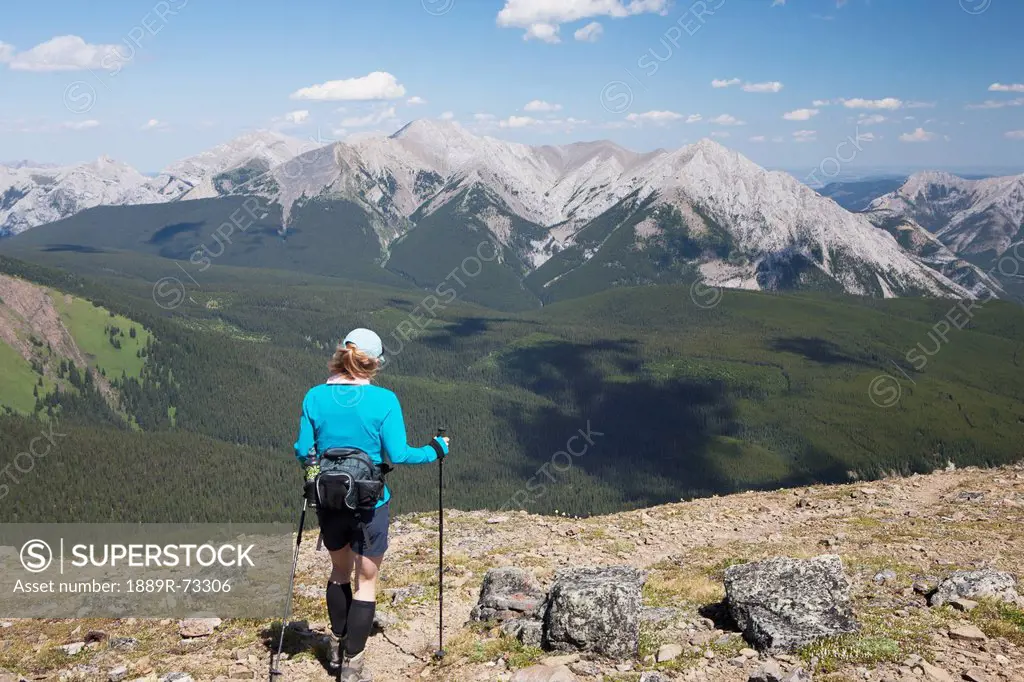 Female Hiker With Hiking Poles Along Ridge Trail With Mountians And Valley In The Distance And Blue Sky And Clouds, Alberta Canada