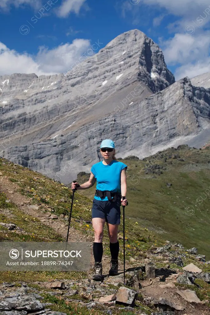 Female Hiker With Hiking Poles On A Mountain Trail With Mountain Peak In The Background And Blue Sky With Clouds, Alberta Canada