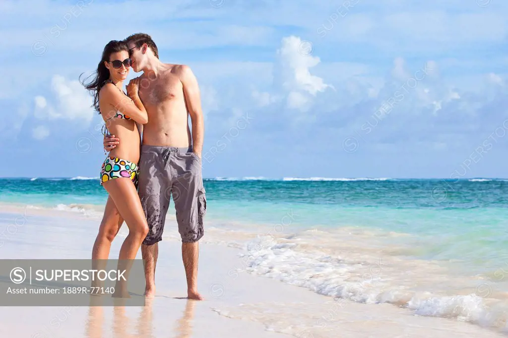 A couple wearing swimsuits stand on the beach at the water´s edge in an embrace, punta cana la altagracia dominican republic