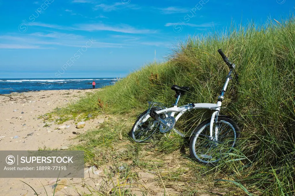 A Bicycle Lays In The Long Grass On The Beach With A Woman Walking In The Distance, Northumberland England