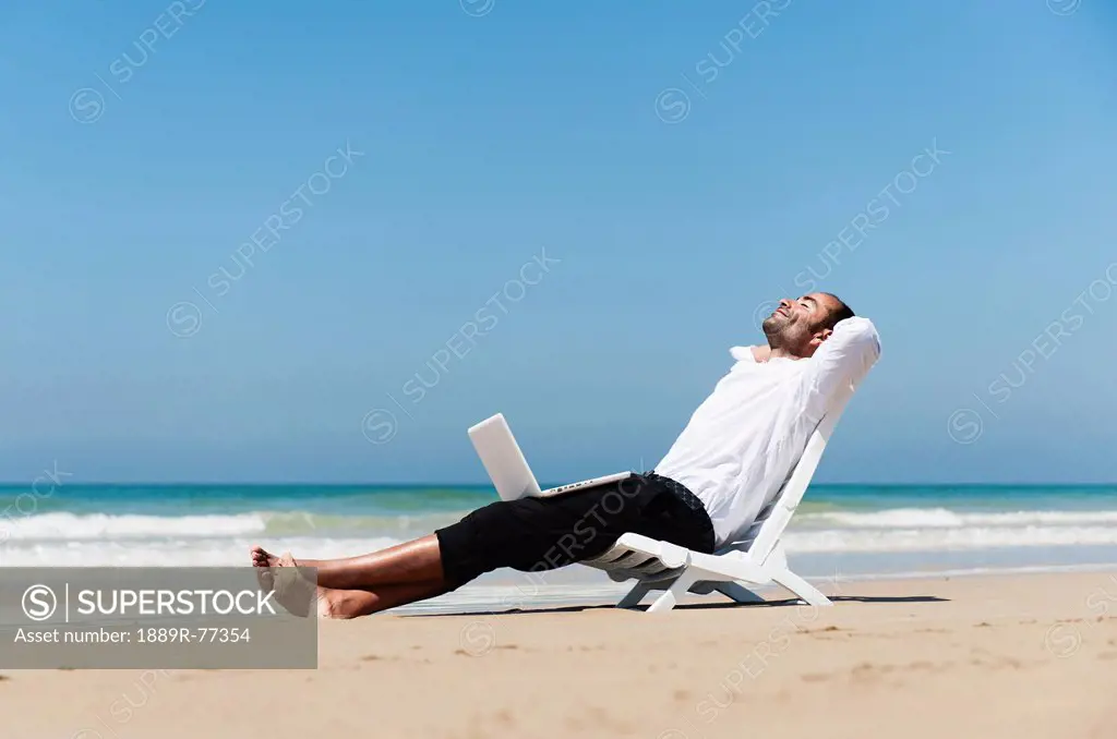 A Businessman Sits On A Beach Chair On The Beach Holding A Laptop Computer On His Lap, Tarifa Cadiz Andalusia Spain