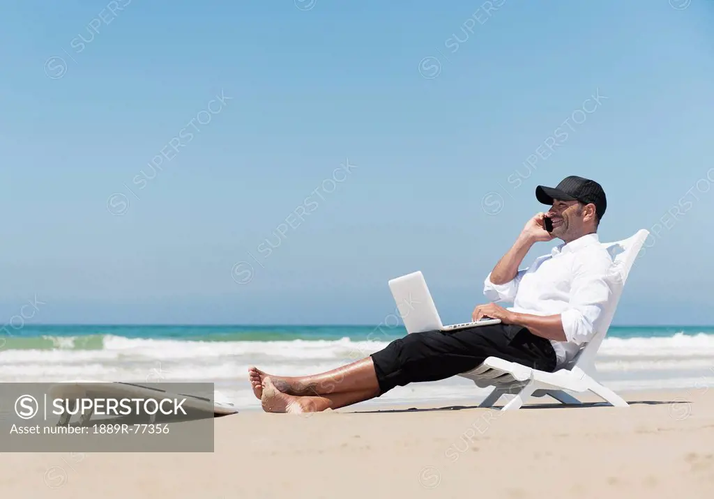 A Businessman Sits On A Beach Chair On The Beach Working On A Laptop Computer And Talking On The Phone With His Surfboard Sitting At His Feet, Tarifa ...