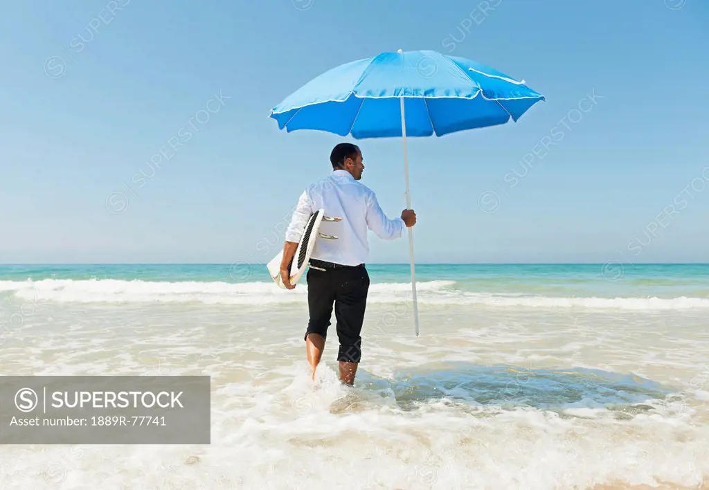 A Businessman On The Beach Holding A Beach Umbrella And Surfboard, Tarifa Cadiz Andalusia Spain