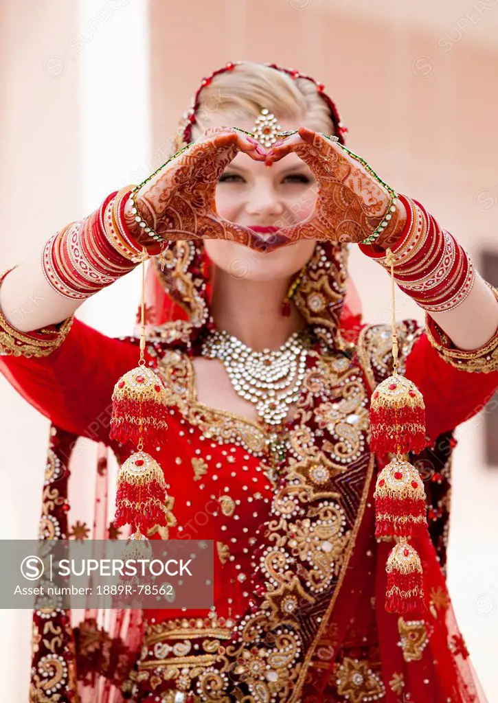 A bride with blond hair wearing a red and gold sari and jewelry holding her mehndi covered hands in a heart shape, ludhiana punjab india