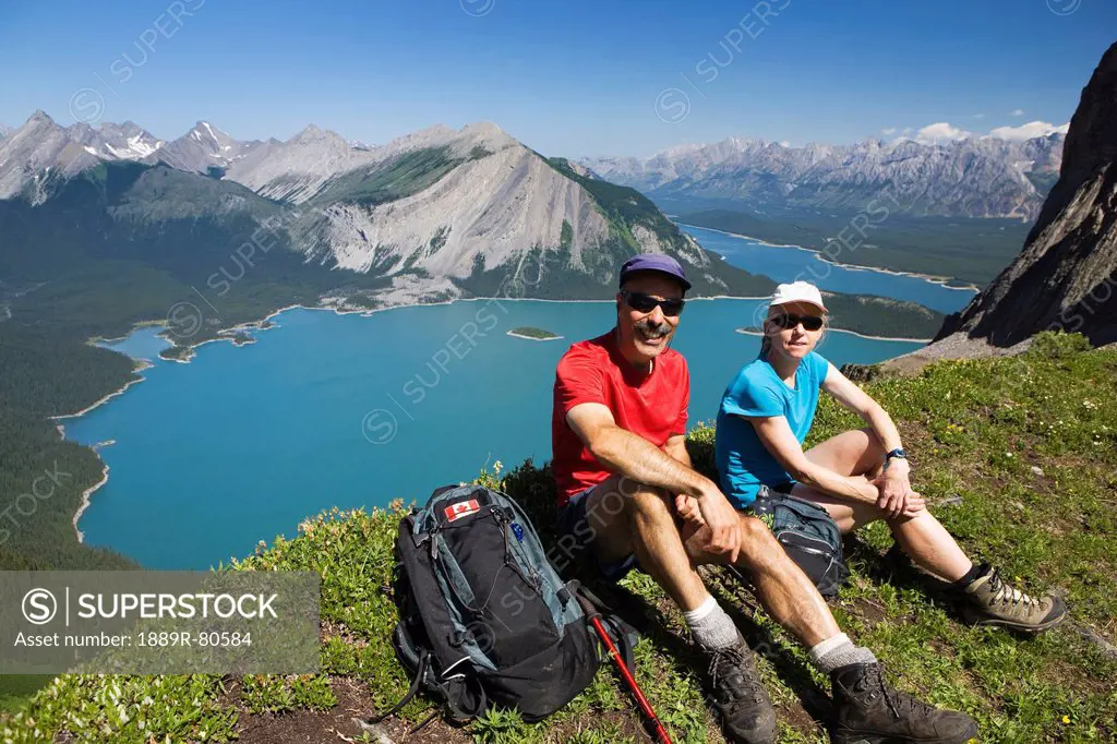 Male and female hiker sitting on top of a mountain ridge overlooking emerald lakes and mountain ranges with blue sky, kananaskis provincial park alber...