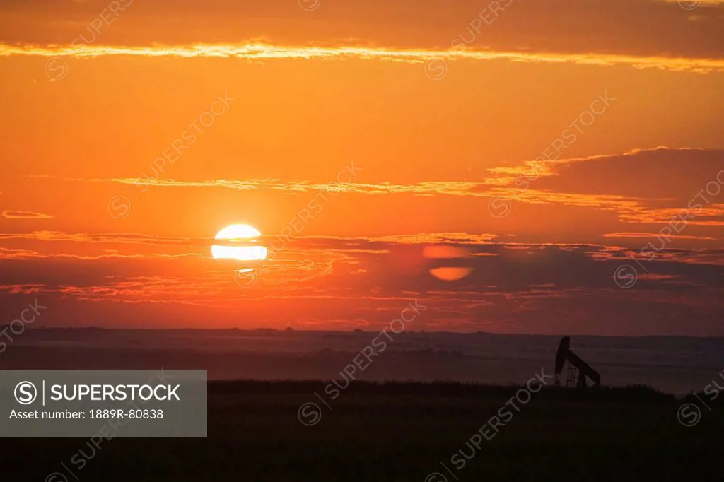 Silhouette of pumpjack in a field at sunrise with orange cast sky and clouds east of airdrie, alberta canada