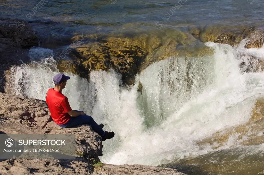 Man sitting on the edge of a cliff looking down on waterfalls at kananaskis provincial park, alberta, canada