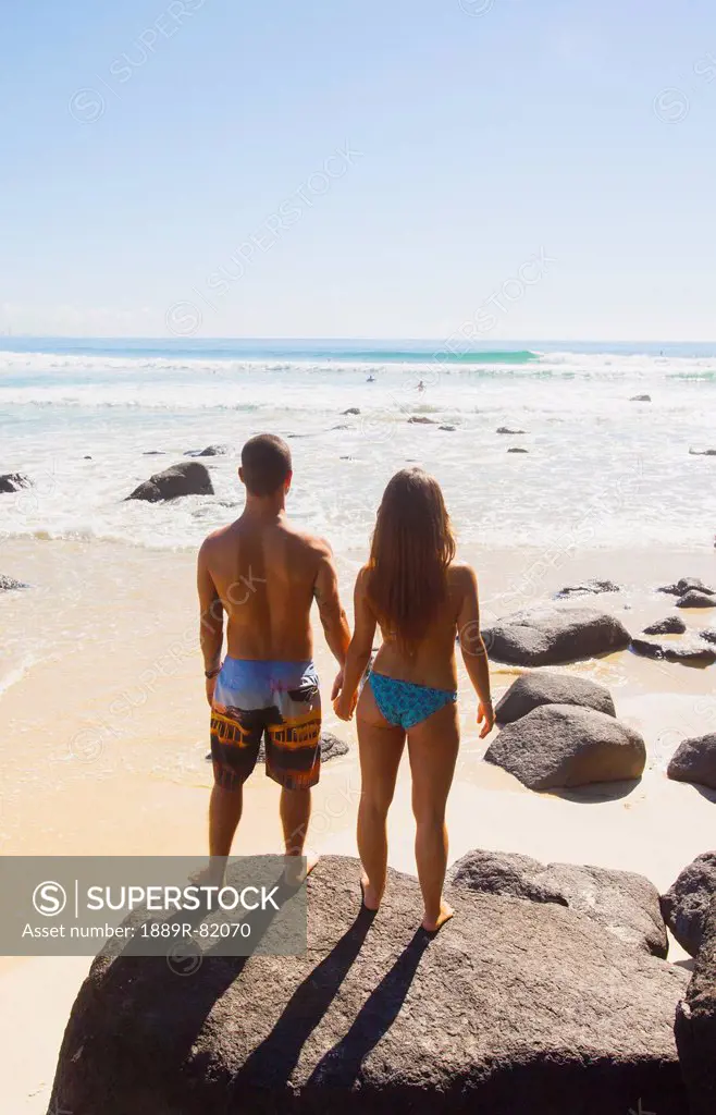 A couple stand in their swimsuits holding hands on a large rock looking out at the ocean, green mount coolangatta gold coast queensland australia