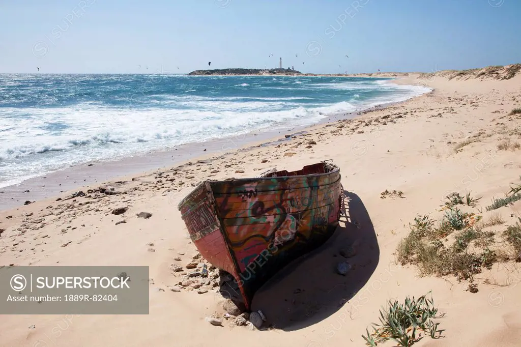 A painted wooden canoe sitting on a beach with the cape trafalgar lighthouse in the background, andalusia, spain