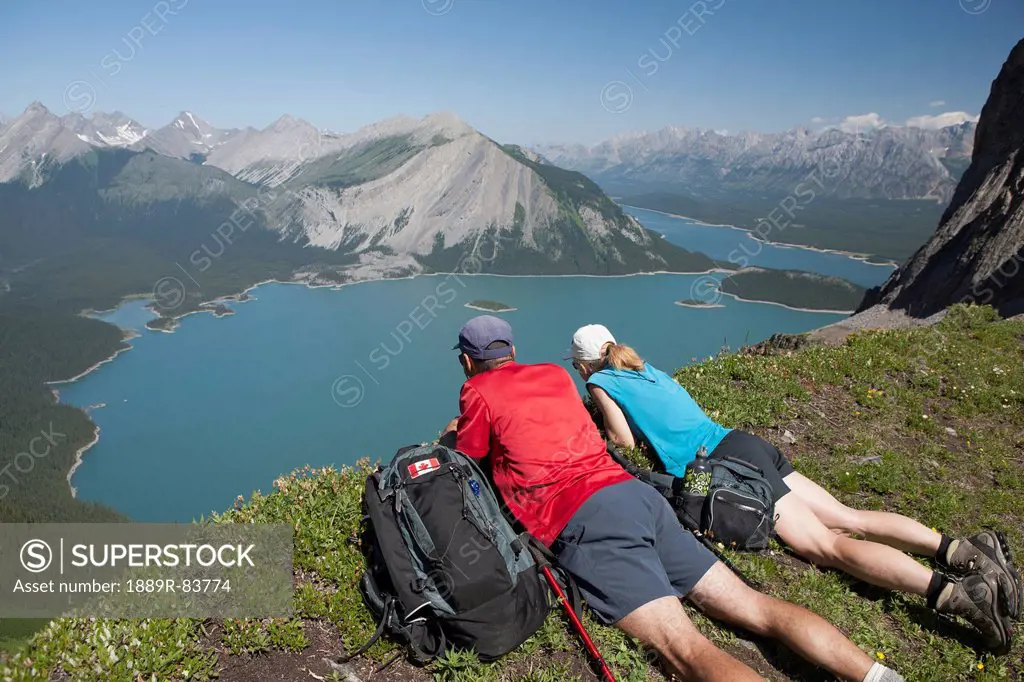 Male And Female Hiker Laying Down On A Mountain Ridge Overlooking An Emerald Lake And Mountains Below With Blue Sky In Kananaskis Provincial Park, Alb...