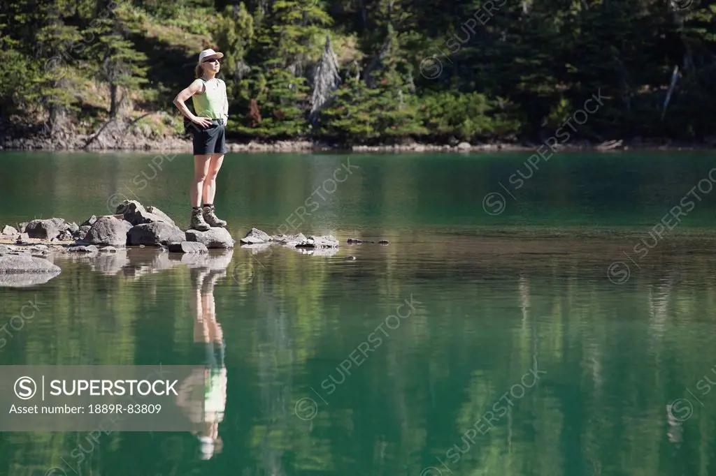 Female Hiker Standing On Rocks At Lake´s Edge Reflecting In Water, Field British Columbia Canada