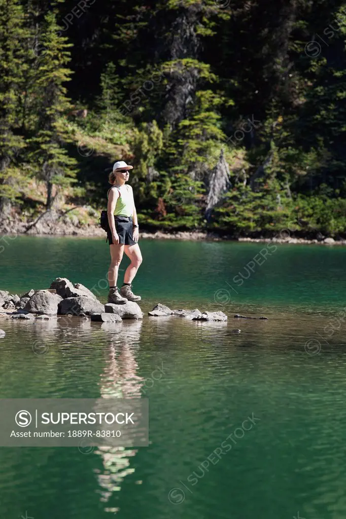 Female Hiker Standing On Rocks At Lake´s Edge Reflecting In Water, Field British Columbia Canada