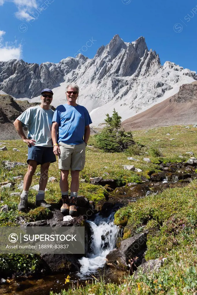 Two Male Hikers Standing Next To A Small Waterfalls In Creek With Mountain Meadows And Mountains In The Backgroud With Blue Sky In Kananaskis Provinci...
