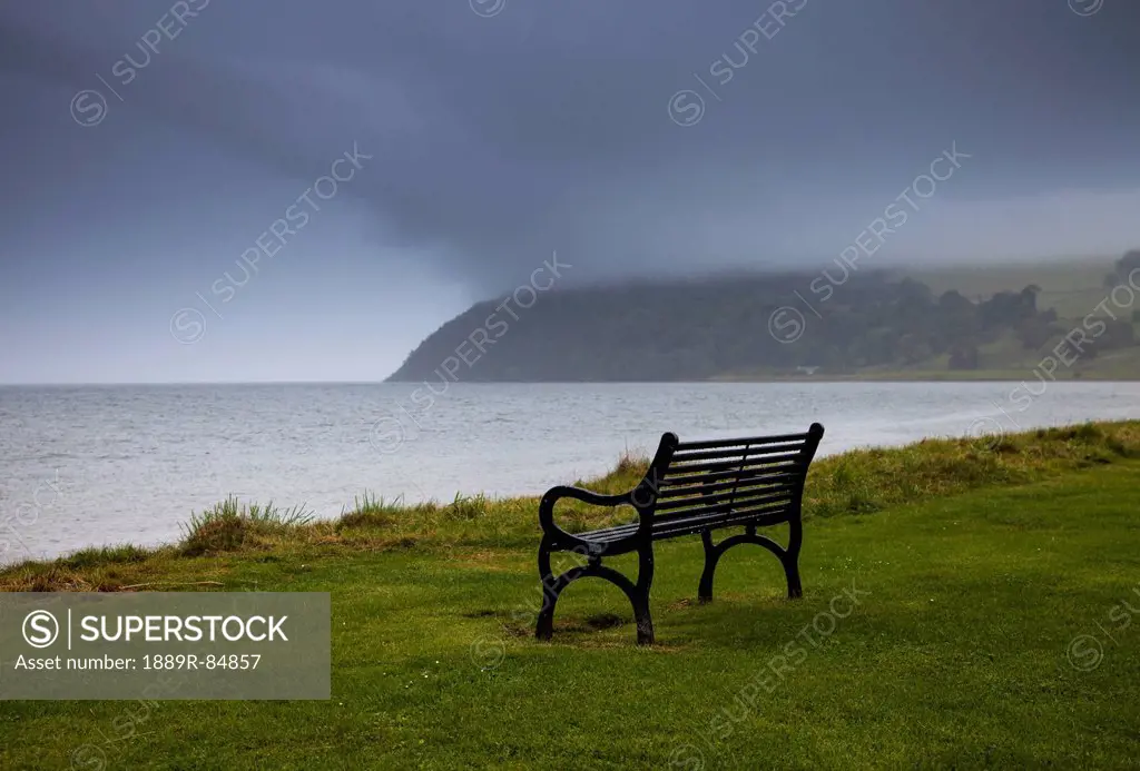 A Bench Sits On The Water´s Edge With Storm Clouds Over The Water, Moray Firth Scotland