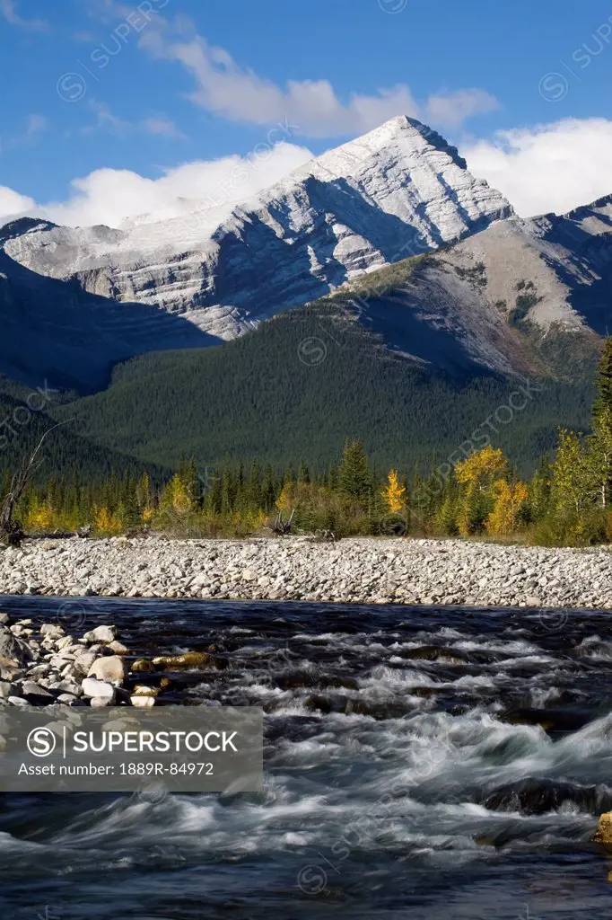 A rushing river with rocky banks and a mountain range with blue sky and clouds in the background in autumn, bragg creek alberta canada
