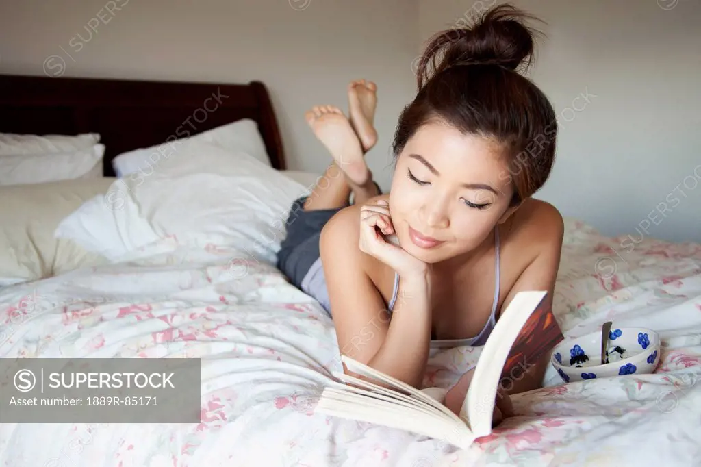 A young woman lays on her bed reading a book, berkeley california united states of america