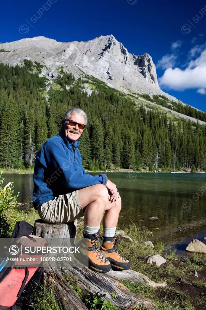 Male Hiker Sitting On A Tree Stump By A Mountain Lake With A Mountain In The Background With Blue Sky And Clouds In Kananaskis Provincial Park, Albert...