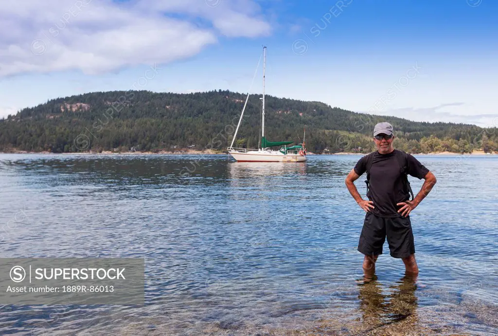 A Senior Man On Shore With His Anchored Sailboat In The Background On Russell Island In The Gulf Islands, British Columbia Canada