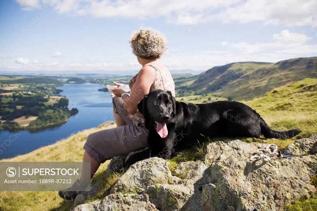 A Woman Sits With Her Dog On A Rock Overlooking A Large Lake And Landscape;Hallin Fell Ullswater Lake District England