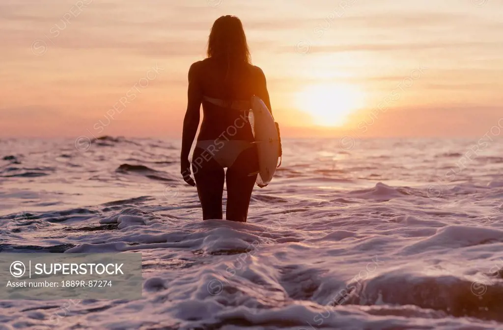 A woman standing in the ocean holding a surfboard at sunset;Canos de meca cadiz andalusia spain
