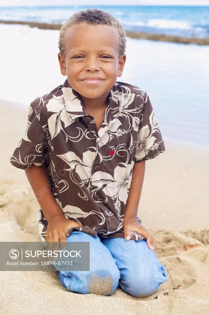 A Young Boy Kneels In The Sand At The Water's Edge;Hawaii United States Of America